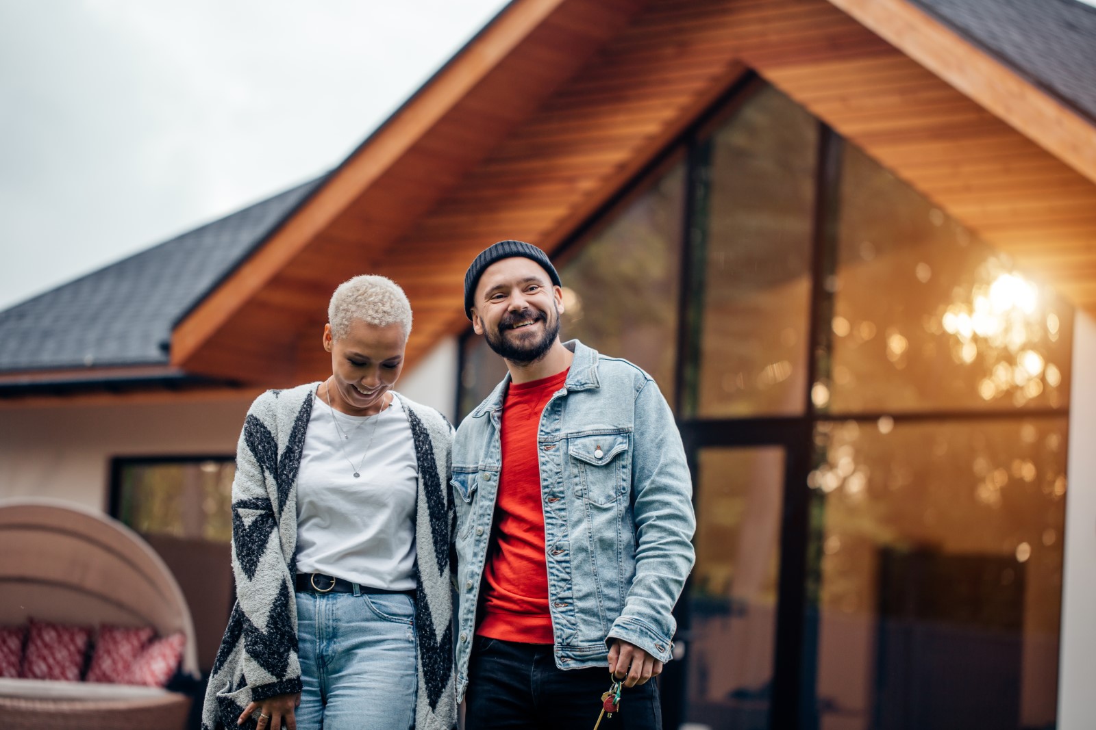 A man and lady wearing a jacket and sweater stand outside of their home in the fall.