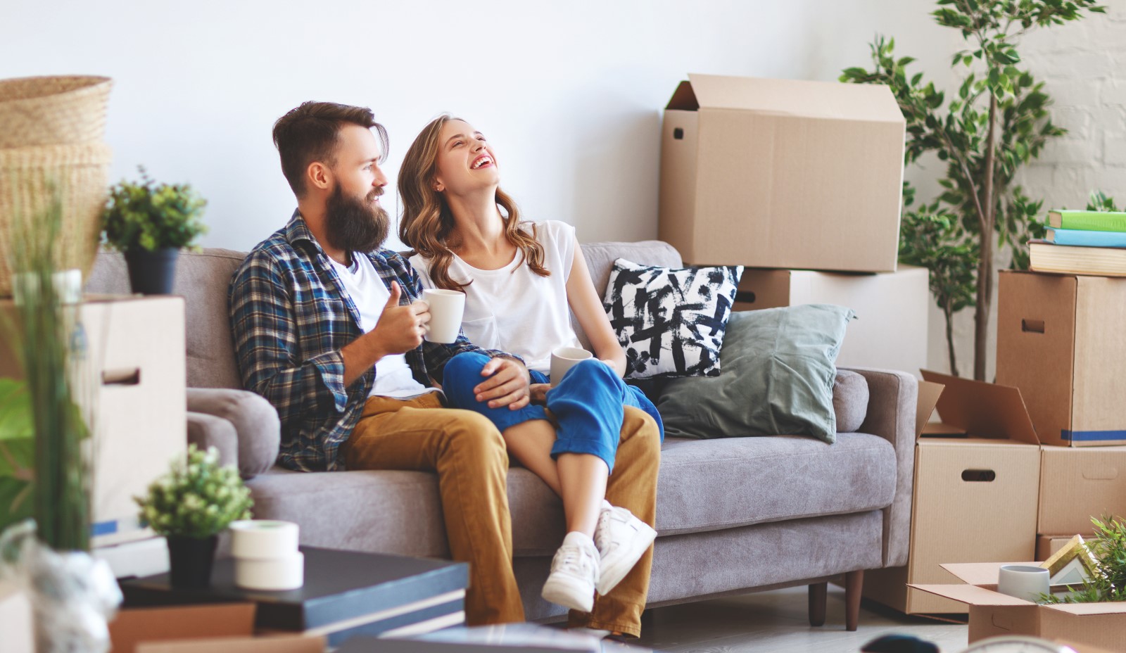 A young couple sits on the couch drinking coffee surrounded by moving boxes.